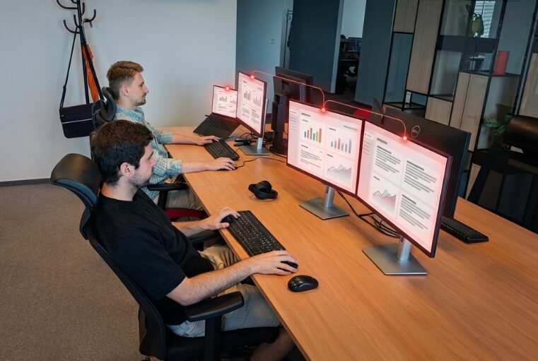 Dvaja kolegovia pracujú na prepojených monitoroch s dashboardmi, medzi ktorými prúdia dáta – tímová spolupráca v multiexperience prostredí